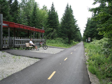 A cyclist sits on a park bench, under a red awning, at a rest stop along the newly paved bike path