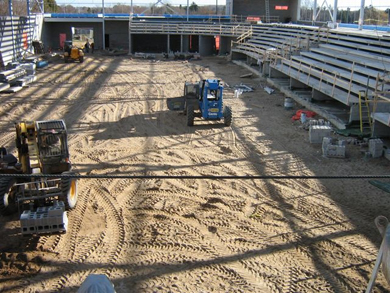 Tandis que les estrades de béton sont déjà en place, des ouvriers déplacent des blocs de béton à l'aide d'élévateurs à fourches sur le sol de terre battue qui deviendra une patinoire