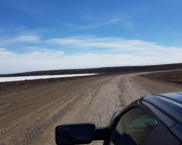 A car on the Inuvik to Tuktoyaktuk highway in the spring of 2018 after it was just opened. The highway is a gravel surface surrounded by local plants and a river.