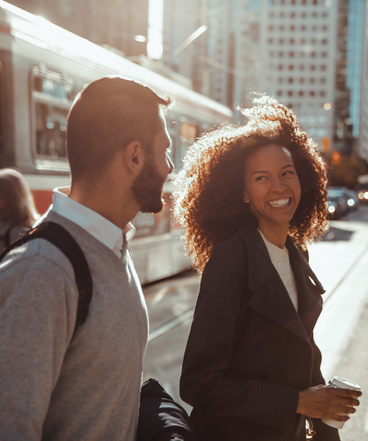 A man and a woman smile looking at each other. The woman is holding a coffee cup and the man is wearing a backpack. In the background, people are walking toward a streetcar in downtown Toronto.
