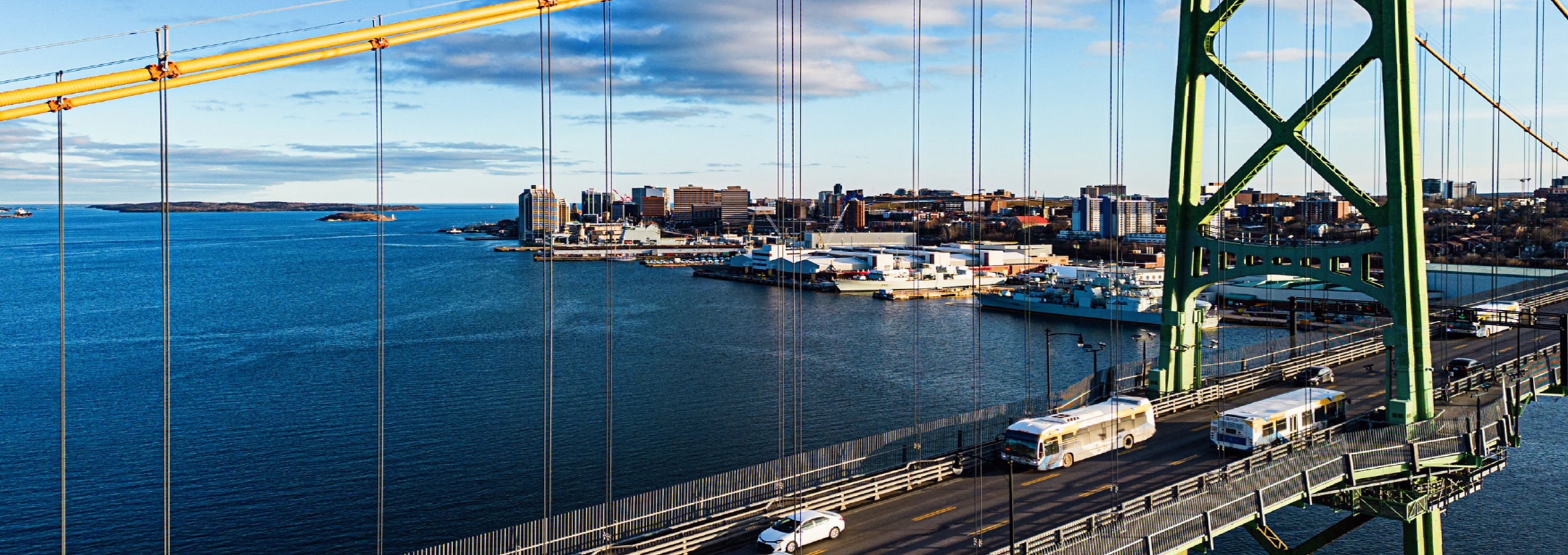 Aerial view of transit buses crossing a suspension bridge overlooking a harbour.