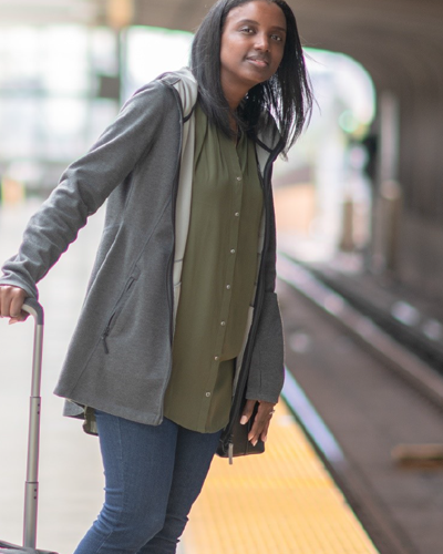 A woman standing on a subway platform leans forward to look for the train. She is holding a suitcase.