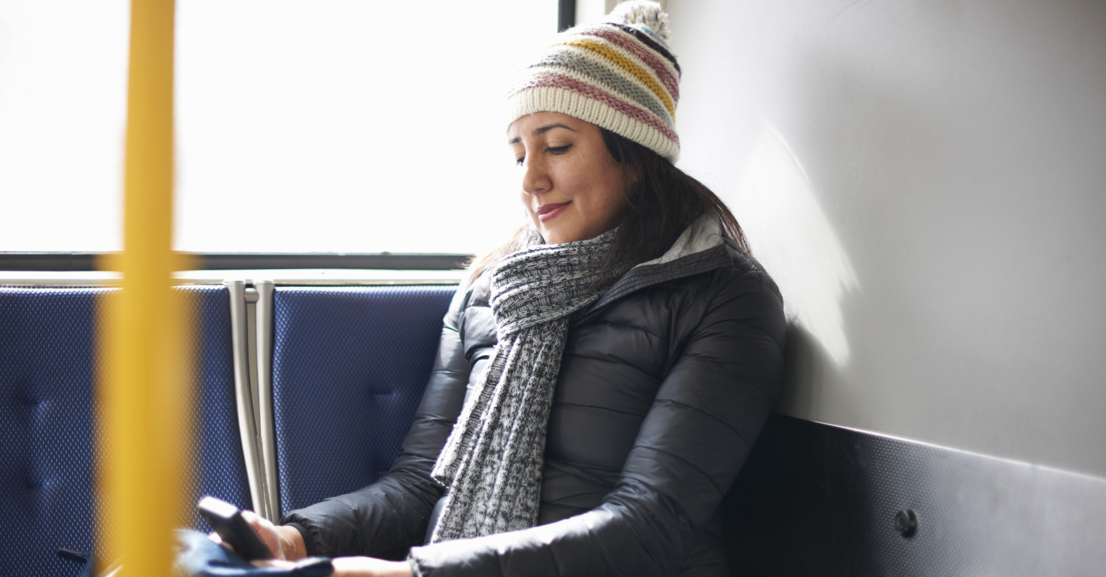 A woman wearing a hat and winter scarf smiles down at her phone while sitting on a bus.